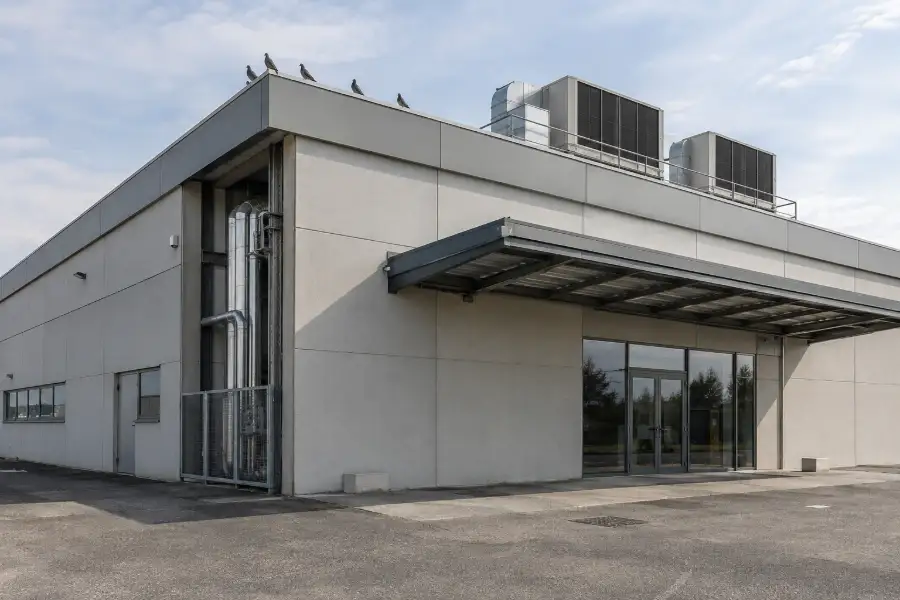Angolo di un edificio commerciale visto dal piazzale, con il lungo cornicione del bordo del tetto, la tettoia metallica sopra l’ingresso vetrato e due unita di trattamento aria sulla copertura piana, con alcuni piccioni posati sul cornicione